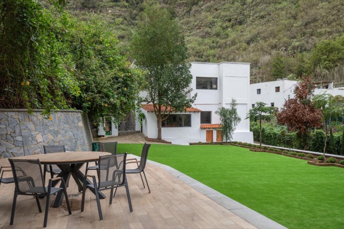 El jardín de la finca, con una mesa al aire libre, la casa blanca y la montaña verde detrás